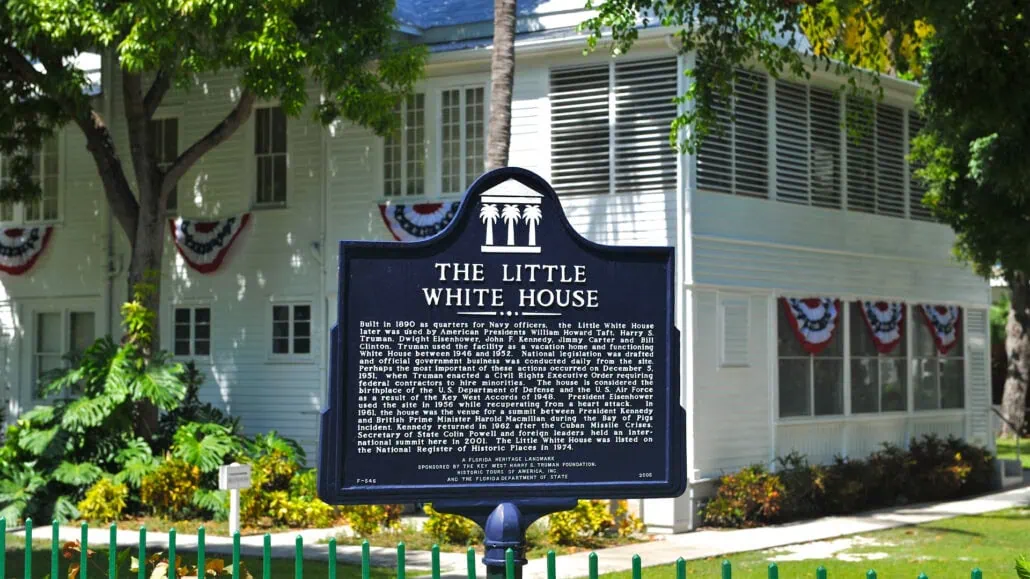A historical marker in front of a white wooden building with red, white, and blue bunting. The sign reads "The Little White House" and provides historical details about the building's origin and significance. This spot is one of the must-do activities in Key West, perfect for cruise passengers exploring local history.