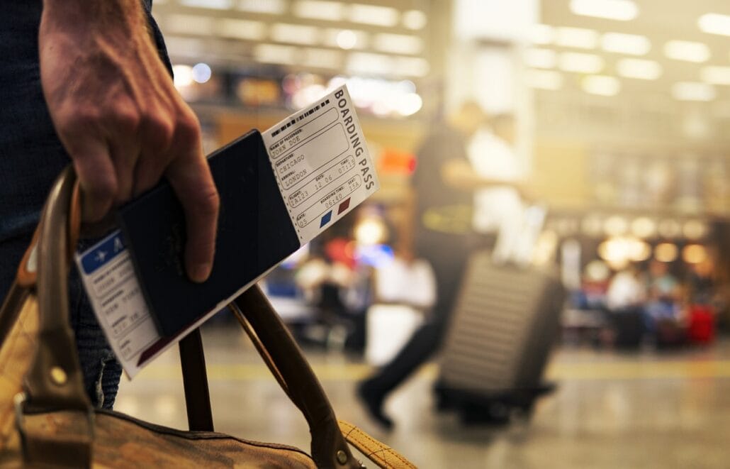 A close-up of a traveler holding a passport and boarding pass in one hand and a brown duffel bag in the other at an airport. The bustling terminal, filled with suitcase-pulling travelers, is abuzz with families sharing travel tips, perfect for cruisers with kids.