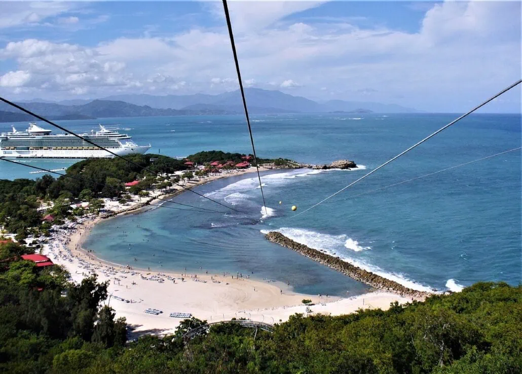 A scenic view of a coastal area with a wide sandy beach, surrounded by green hills and calm blue sea. Two family cruises are docked in the distance. Zipline cables stretch across the image, suggesting a nearby ziplining activity that could involve kids in an adventurous day.