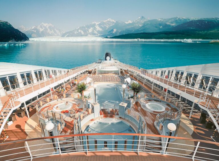 The image shows the deck of a cruise ship with multiple pools and loungers. It is sailing in clear blue water, surrounded by snow-capped mountains and glaciers in the background under a clear sky.