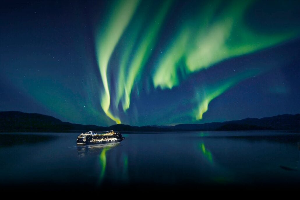 A cruise ship is illuminated at night on calm waters, with the Northern Lights glowing green above in the clear night sky. Hills are visible on the horizon, and the lights are reflected in the water.
