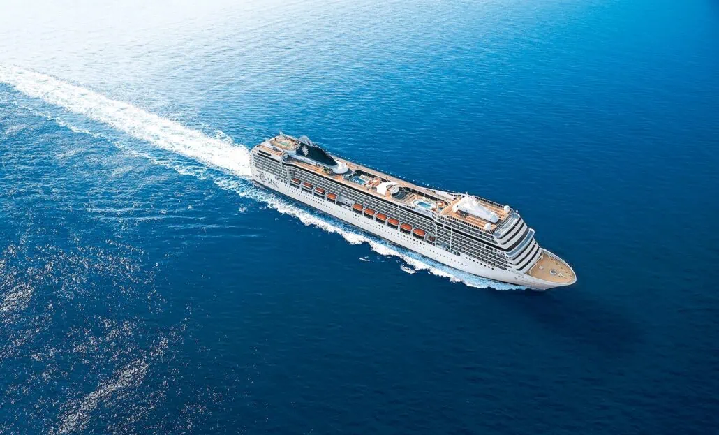 A large cruise ship travels through a vast expanse of deep blue ocean, leaving a white wake behind it. The ship's multiple decks are visible, and the clear sky is reflected in the water.