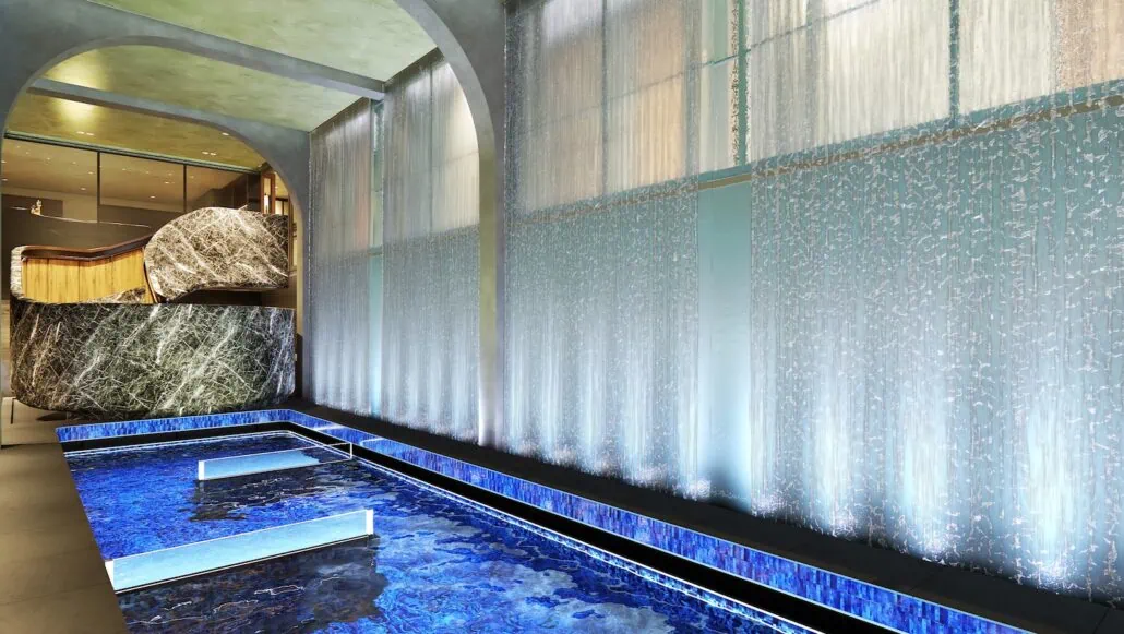 A modern indoor space aboard one of Norwegian's ships features a blue lit water installation with cascading water walls. Large marble structures and archways create an elegant and serene atmosphere, reminiscent of an upgraded spa.