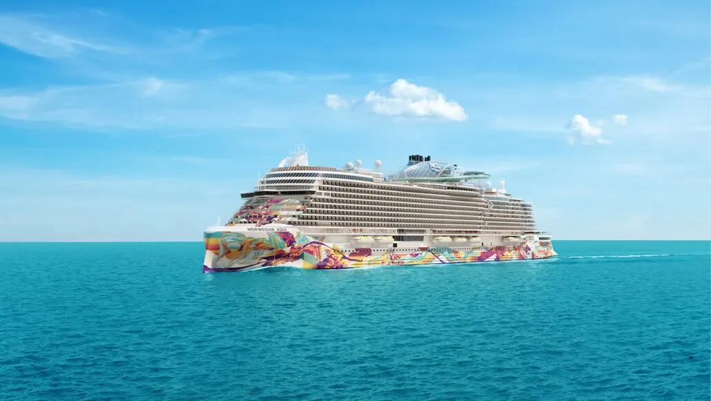 A large, multi-story Norwegian cruise ship with colorful artistic designs on the bow sails through calm, blue ocean waters under a clear sky with a few clouds, promising relaxation and luxury at its onboard spa.