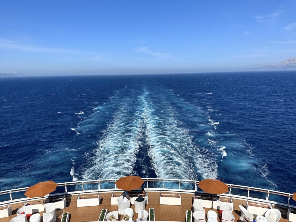 View from the aft balcony cabin of a cruise ship shows its wake trailing across the vast blue ocean. The upper deck features several seating areas with white chairs and brown umbrellas. There's a clear horizon under the sunny sky, highlighting the pros of an upgrade to this view.