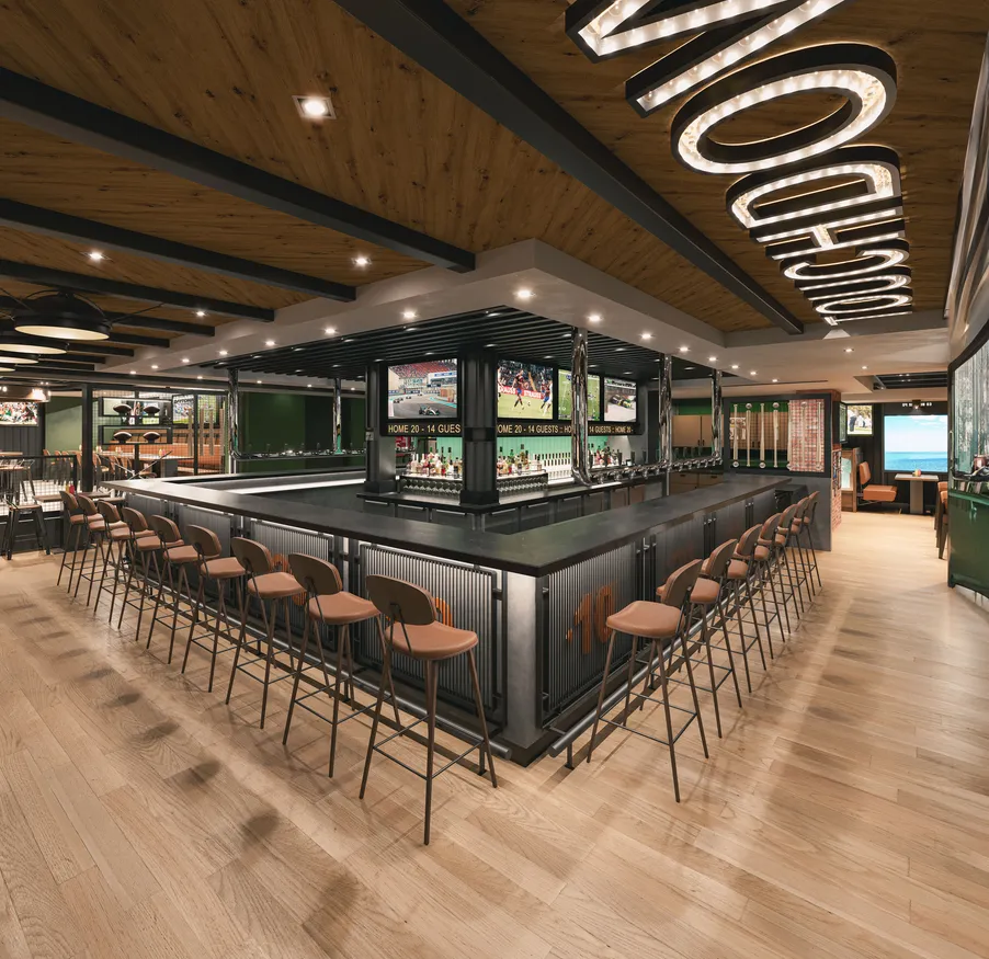 A modern, spacious lounge reminiscent of MSC World America features a central rectangular bar surrounded by high stools. The ceiling boasts wooden panels and large illuminated letters, while multiple TV screens display sports games above the light wood floor.