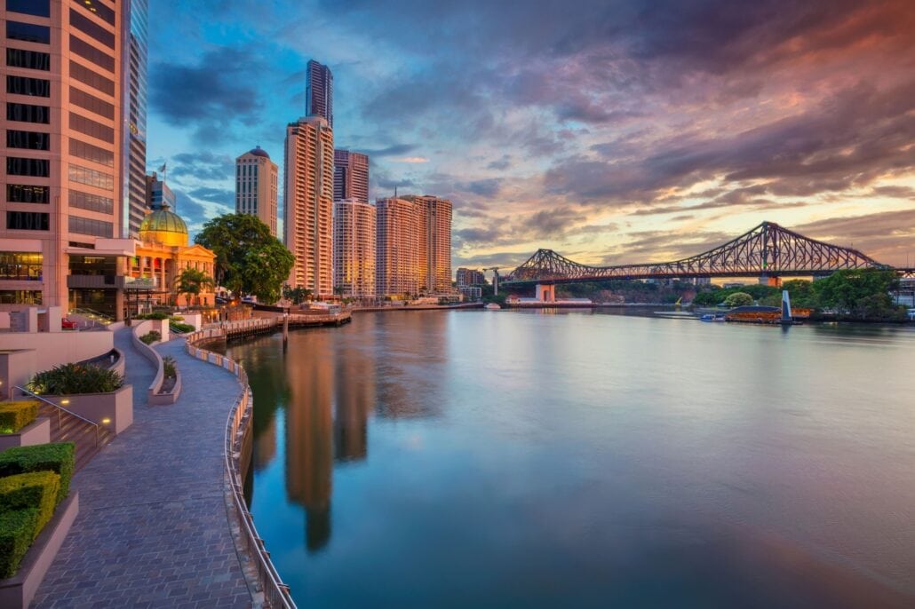 Riverside view of a cityscape at sunset, featuring tall buildings, a river, and a large bridge. The sky is partly cloudy with warm colors reflecting on the water. Alongside the river winds a curved pathway bordered by greenery and modern architecture, showcasing vibrant Australian offerings.