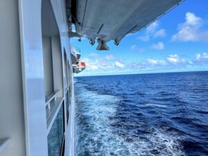 View from a ship's deck at Carnival Cove reveals the ocean and a clear blue sky. Lifeboats are mounted above the deck, and a trail of white foam follows behind, almost as if inviting passengers to consider a cruise upgrade or relax on their private balcony.