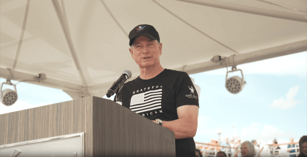 A man in a black cap and T-shirt stands at a wooden podium with a microphone, delivering an auto draft speech outdoors under a large white canopy. The sky is blue with a few clouds drifting by in the background.