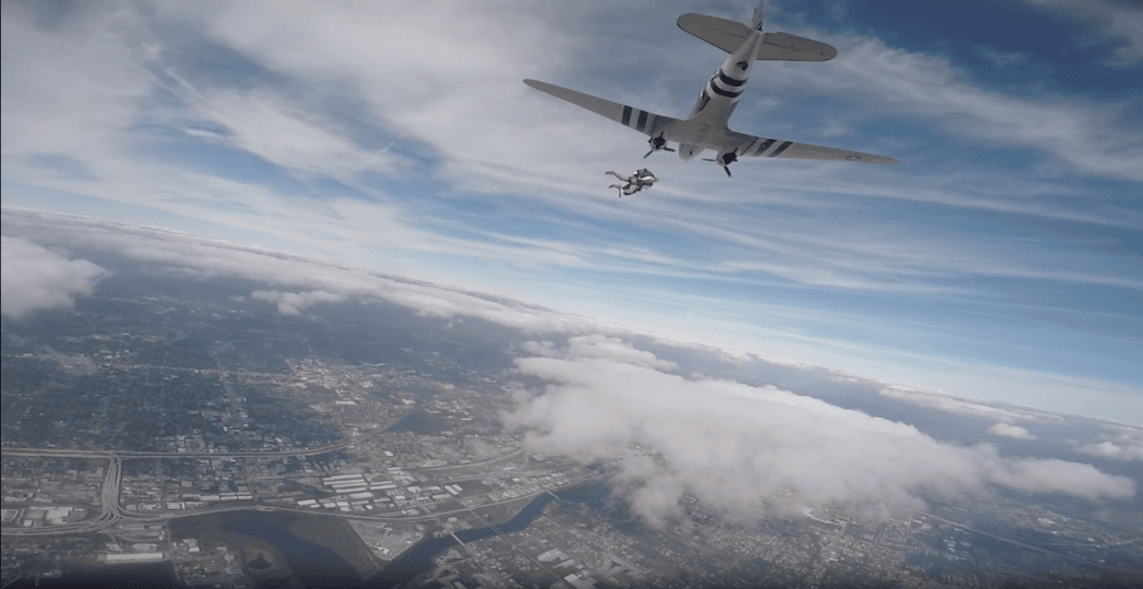 A skydiver leaps from an aircraft adorned with distinctive black and white stripes, soaring over a cityscape like an auto draft in the wind. Below, the ground is a tapestry of urban development, rivers, and scattered clouds beneath a mostly clear sky.