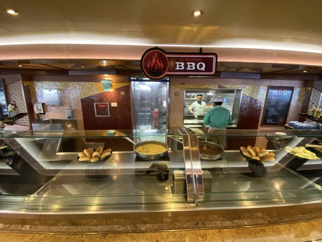 A BBQ restaurant counter with various dining options displayed, including bread rolls, rice, and meat dishes. Two staff members are in the kitchen area behind the counter. A sign above reads "Old Fashioned BBQ," making it one of the must-visit places to eat on your Carnival Freedom journey.