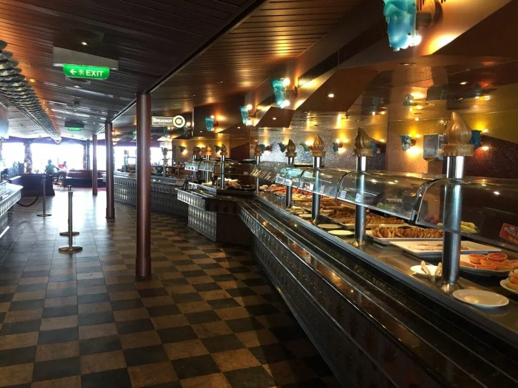 The buffet area on the Carnival Freedom features a long counter of dining options under glass covers. With a wooden ceiling adorned with light fixtures and a checkered floor. An exit sign is visible in the upper left corner, guiding guests to other enticing places to eat on board.