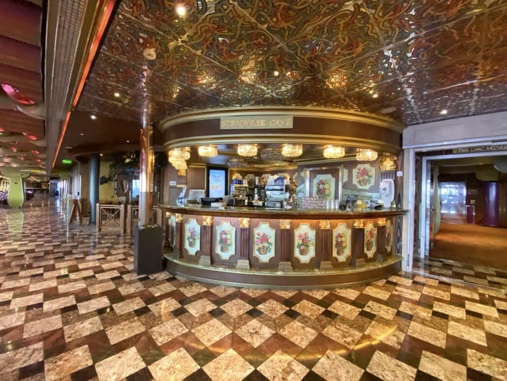 A vintage-style café aboard Carnival Freedom, with ornate decorations, a patterned ceiling, and a tiled floor. As one of the enticing dining options, the counter displays various items beneath an illuminated sign. This inviting place to eat exudes warmth and charm.