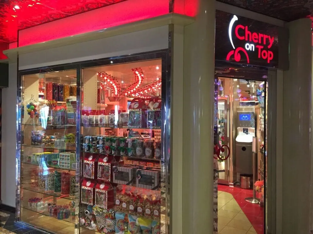 Cherry on Top," a brightly lit candy store reminiscent of Carnival Freedom, boasts a neon sign. The storefront tempts with various candies, packaged sweets, and colorful lollipops. Inside, the red decor and warm lighting create a delightful place to eat sweet treats.
