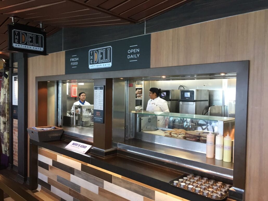 A deli counter with two staff members showcases a variety of sandwiches. The sign reads "The Deli Artisan Eats" under "Fresh Food Open Daily." Several sauce bottles are neatly arranged, making it a delightful place to eat reminiscent of the inviting atmosphere aboard Carnival Cruise Line.