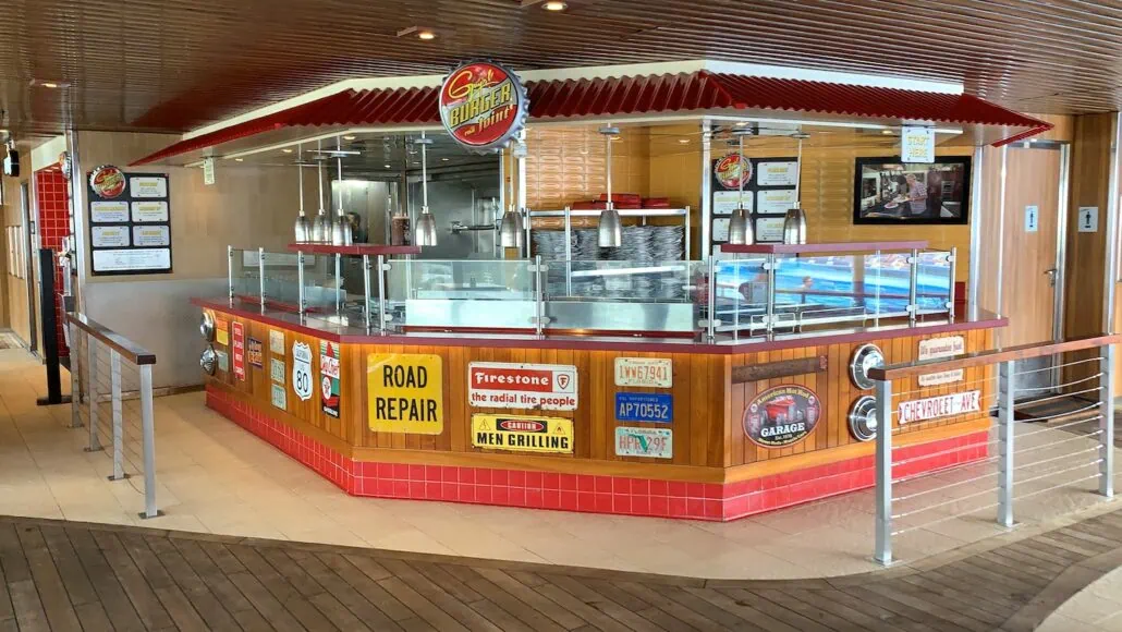 A casual outdoor food stand with a retro American diner theme, located among popular places to eat near the Carnival Freedom dock. It features a striped red and yellow awning, vintage advertisement signs, and metal accents. The counter is enclosed with clear glass panels, and pendant lights hang overhead.
