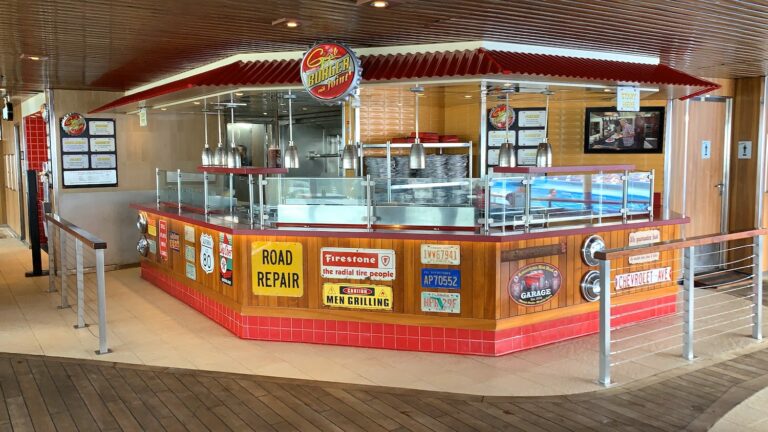 A casual outdoor food stand with a retro American diner theme, located among popular places to eat near the Carnival Freedom dock. It features a striped red and yellow awning, vintage advertisement signs, and metal accents. The counter is enclosed with clear glass panels, and pendant lights hang overhead.