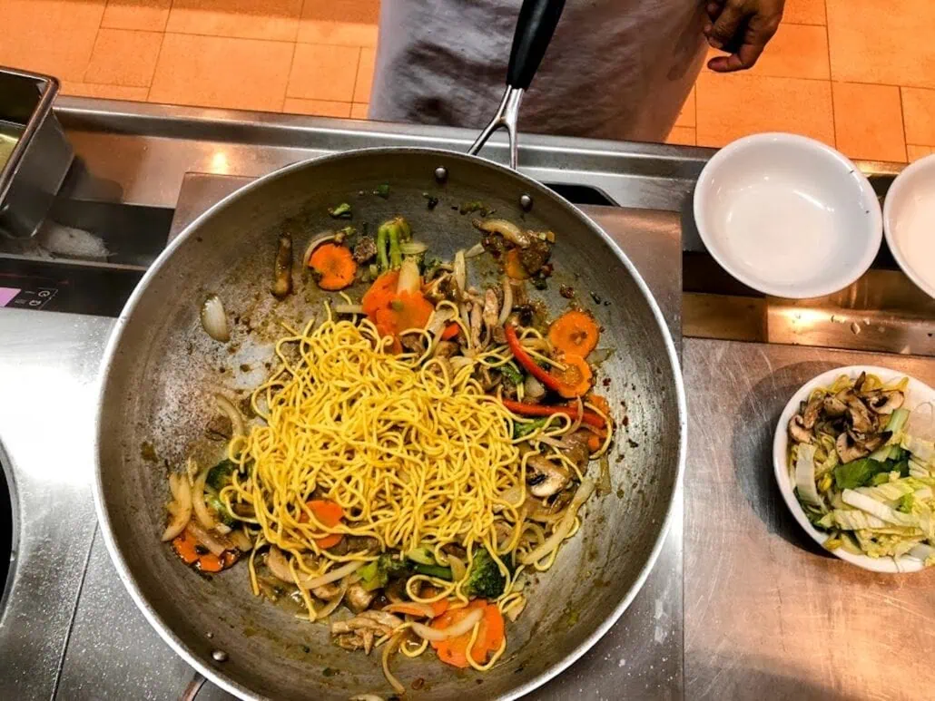 A wok on a stove contains stir-fried noodles with vegetables like carrots, broccoli, and mushrooms. A person stands nearby in a kitchen reminiscent of the diverse dining options found aboard Carnival Freedom. A bowl of additional chopped vegetables and empty bowls are on the counter.