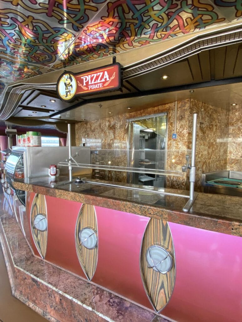The empty pizza counter at "Pizza Pirate" on Carnival Freedom showcases a colorful, patterned ceiling. A glass display case and metallic counter with a marble-like top are visible, hinting at one of the many dining options available onboard. Kitchen equipment hums quietly in the background.