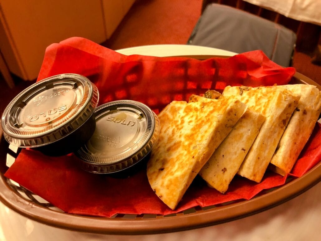 A basket of quesadilla slices, lined on red paper, invites you to explore one of the delightful dining options aboard Carnival Freedom. Accompanied by two small black sauce containers with silver lids, it rests on a table with a pleasantly blurred background.