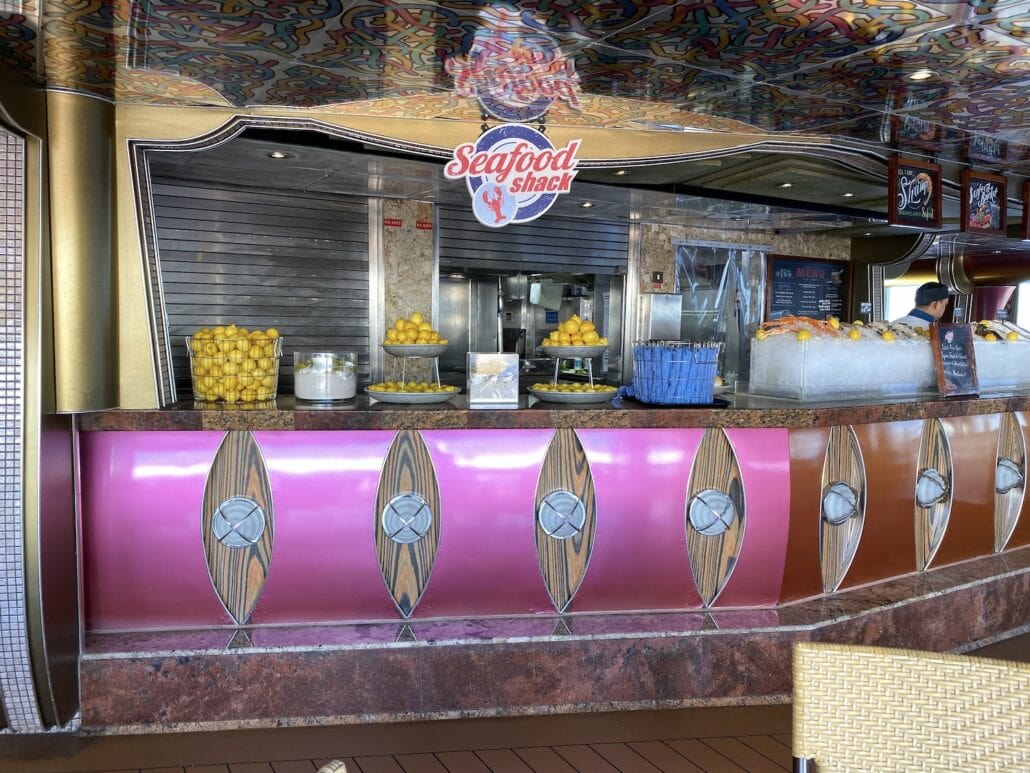 A seafood counter titled "Seafood Shack" on Carnival Freedom offers enticing dining options. Adorned with lemons and a basket of citrus, it showcases seafood nestled in ice trays. The staff member adds a lively touch, while the colorful, patterned ceiling completes this inviting place to eat.