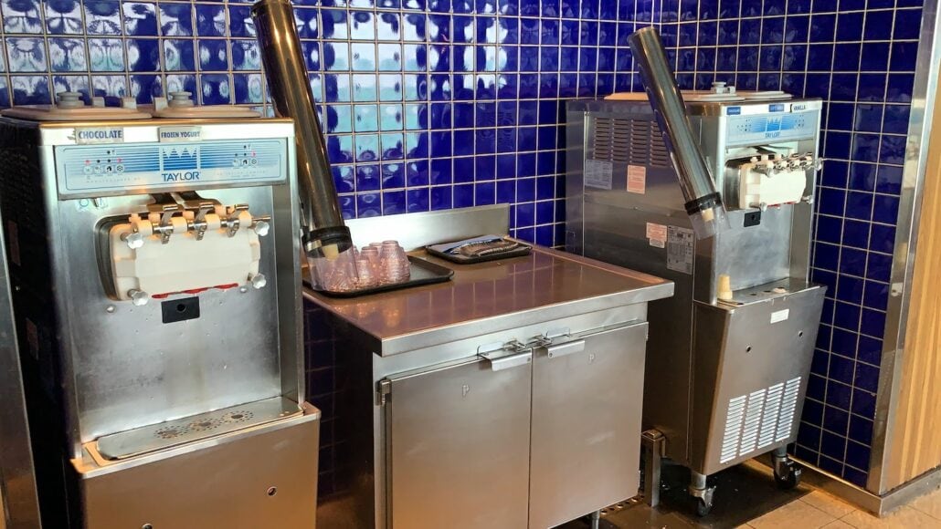 Two soft-serve ice cream machines grace a wall adorned with blue tiles on the Carnival Freedom. A tray of clear plastic cups rests on the stainless steel counter between them, perfect for a delightful treat while enjoying Carnival Cruise Dining. Cones are conveniently dispensed from each machine.