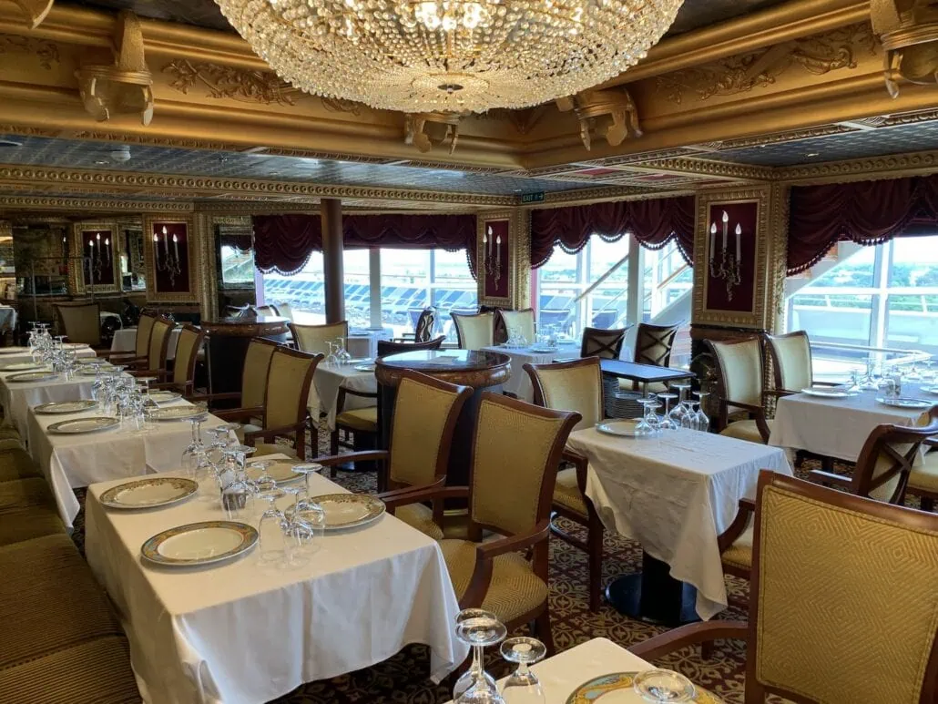 A luxurious dining room aboard the Carnival Freedom features elegantly set tables and chandeliers. Plush chairs and ornate details complement the white tablecloths, plates, and glassware. Large windows with draped curtains offer a scenic view, adding to the refined dining options.