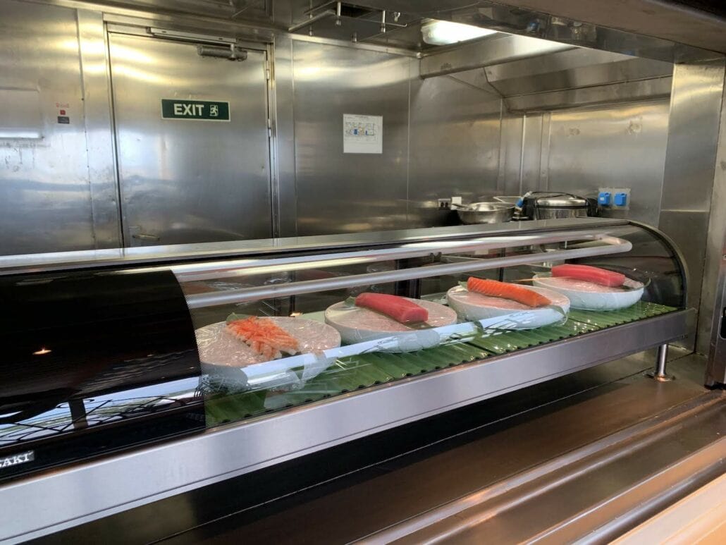 A vibrant sushi counter display features five plates of sushi on a conveyor, each wrapped in plastic. The background reveals a bustling stainless steel kitchen with an exit sign and some equipment, highlighting the diverse dining options reminiscent of places to eat on Carnival Freedom.