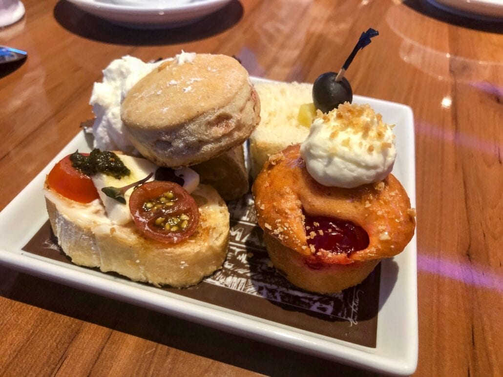 On a wooden table aboard the Carnival Freedom, a plate of assorted pastries and appetizers entices. Items include a cherry-topped muffin, tomato and cheese bruschetta, a scone with cream, a slice of cake, and a small sandwich garnished with a black olive—a delightful scene for those seeking places to eat.