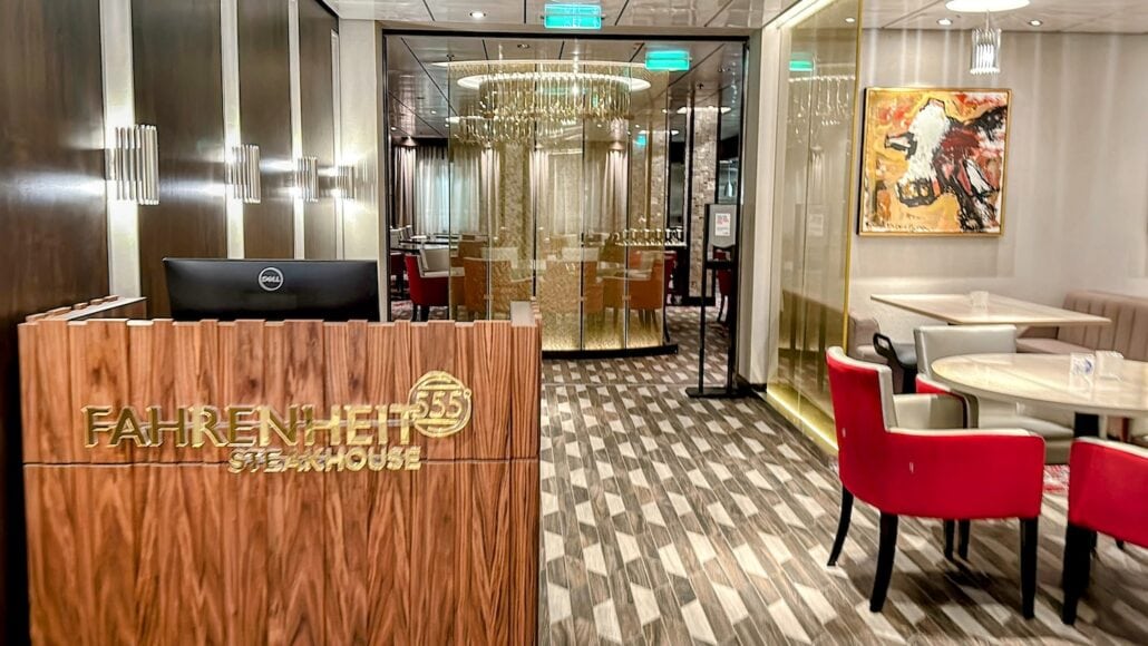 The image shows the interior of a restaurant named Fahrenheit 555 Steakhouse. It features a wooden reception desk, red and beige chairs, a glass partition with a chandelier, and a framed abstract painting on the wall.
