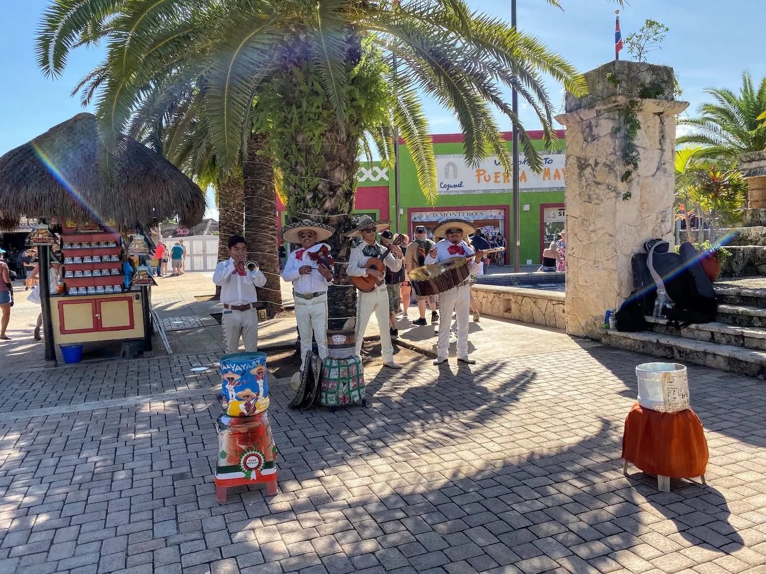 A group of musicians in traditional attire perform under a palm tree on a sunny day at one of the lively Caribbean cruise ports. They are surrounded by a stone structure, a small vendor stand, and a plaza area with shops in the background. A decorative sign and hat are placed in front.