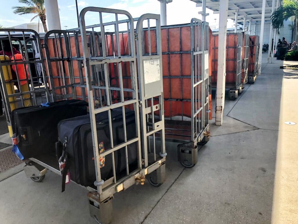 Three luggage carts are positioned outside a building. Two carts are mostly empty with orange padded interiors visible, while one cart holds several black suitcases. A palm tree is in the background, and it appears to be a sunny day at the bustling Florida port, amidst recent labor union discussions.