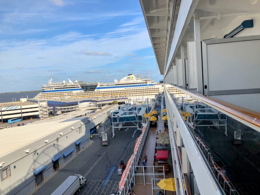 A view from the side of a cruise ship reveals a bustling dock and another majestic cruise vessel in the distance. The sky is mostly clear with a few clouds. A baggage handler moves swiftly among parked vehicles, cargo, and occasional umbrellas, embodying the heartbeat of this seafaring world.