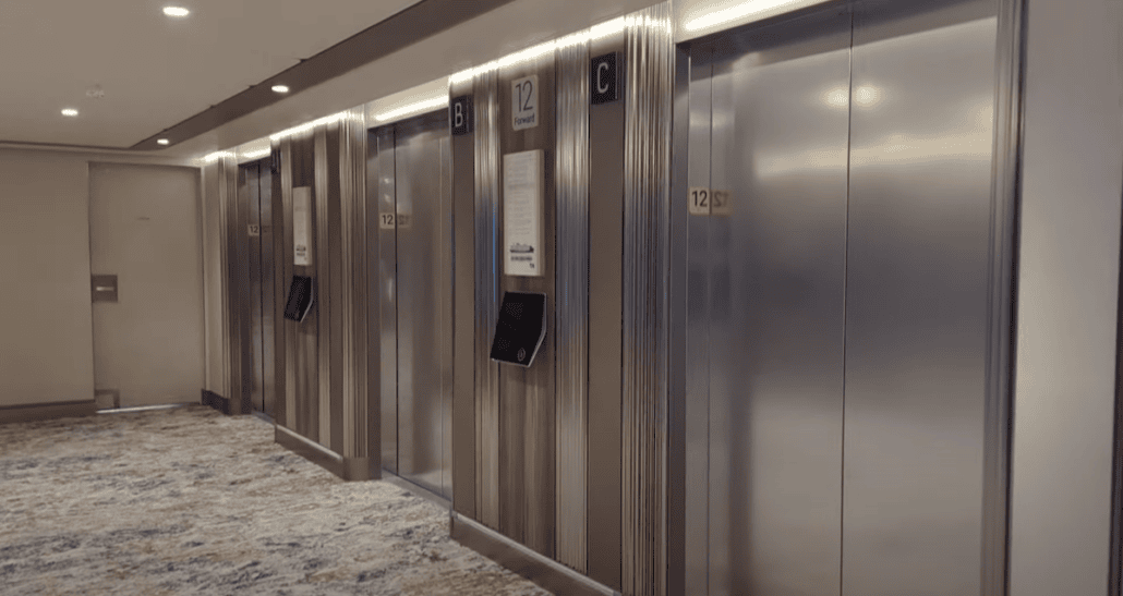 A modern hallway resembles a sleek cruise ship corridor, featuring four elevator doors labeled A, B, C, and D. Each door has a digital screen and a panel displaying the number 12 for passengers. The area is carpeted and softly lit with recessed lighting.