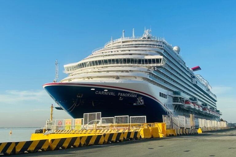 The Carnival Panorama, proudly representing Carnival Cruise Line, docked at the port during its inaugural visit. The ship's white facade and dark blue hull stand tall against a clear blue sky, with safety barriers lining the dock as it brings excitement and grandeur to its guests.