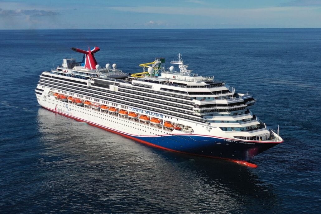 A large Carnival cruise ship sails on a calm ocean under a clear blue sky. The vessel features multiple decks, a distinctive red and blue funnel, and various water slides and amenities visible on the top deck, showcasing its vibrant Australian offerings.