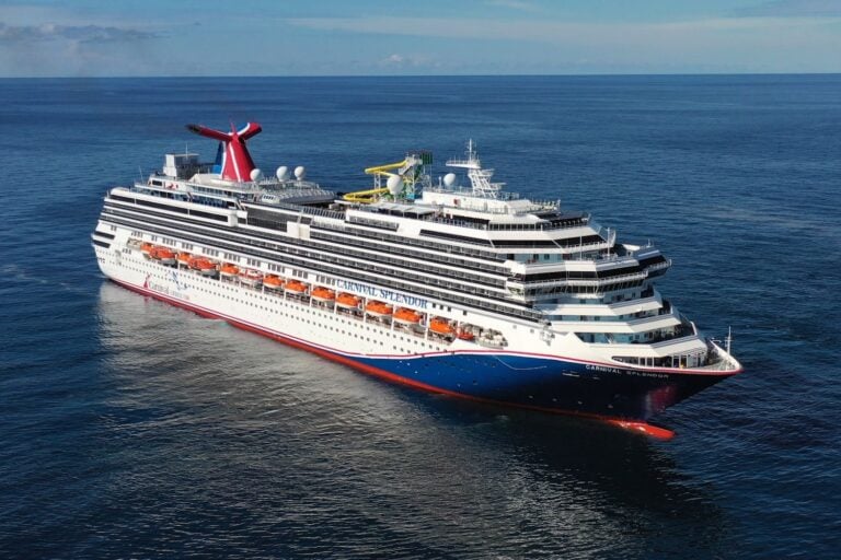 A large Carnival cruise ship sails on a calm ocean under a clear blue sky. The vessel features multiple decks, a distinctive red and blue funnel, and various water slides and amenities visible on the top deck, showcasing its vibrant Australian offerings.