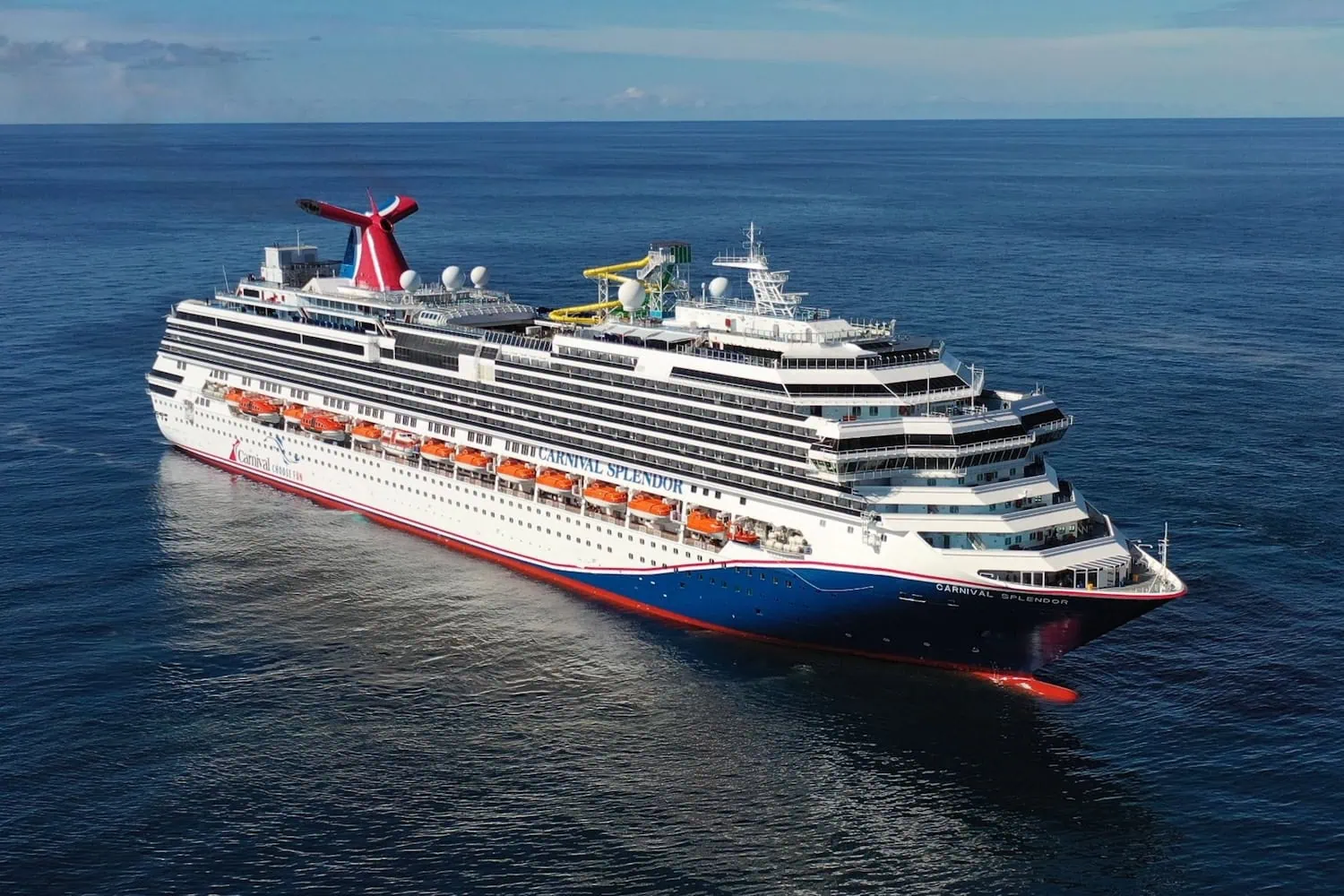 A large Carnival cruise ship sails on a calm ocean under a clear blue sky. The vessel features multiple decks, a distinctive red and blue funnel, and various water slides and amenities visible on the top deck, showcasing its vibrant Australian offerings.