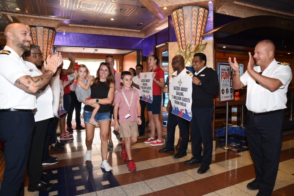 A group of people, including children and adults, walk through a corridor while being applauded by ship crew members in uniforms. The Carnival Cruise Line crew holds signs welcoming them aboard, mentioning activities for young guests as they embark on their New Orleans adventure.