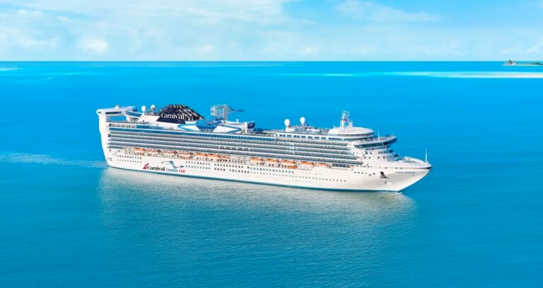 A large white cruise ship from the Carnival Cruise Line sails in a calm, bright turquoise sea under a clear blue sky. With multiple decks and lifeboats visible, this vessel showcases the elegance of Carnival ships as a distant shoreline fades gently in the background.