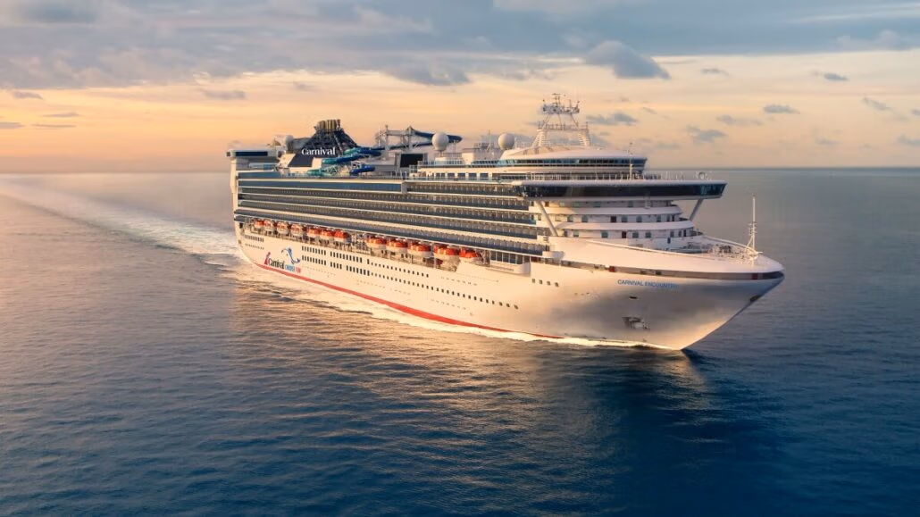 A large Carnival Cruise Line ship sails on calm blue waters under a clear sky. The ship boasts multiple decks, with lifeboats visible on the side. A wake trails behind the vessel, showcasing its graceful motion across the ocean.
