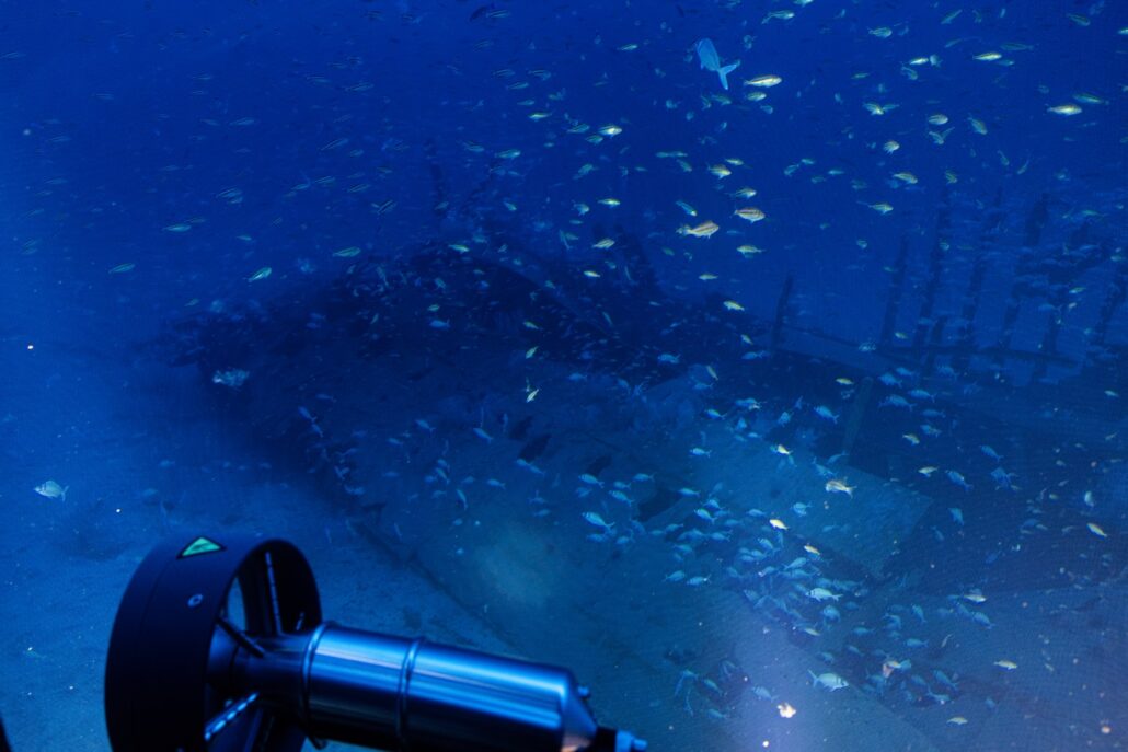 A WWI shipwreck looms in the distance, partially visible under the deep blue waters, where numerous small fish dart playfully. In the foreground on the left side, a metal underwater scooter rests, hinting at a recent cruise expedition exploring this century-old maritime relic.