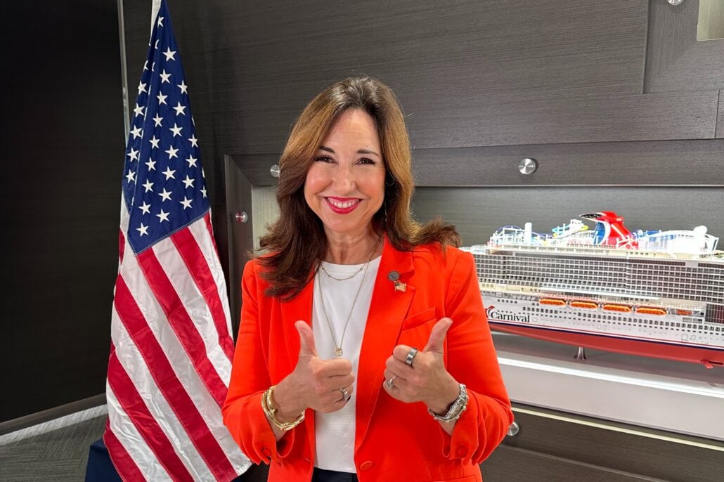 A woman in an orange blazer, representing Carnival Cruise Line, gives two thumbs up with a smile as she stands before an American flag and model cruise ship. Wearing a white top, she is framed by a wood-paneled wall backdrop.