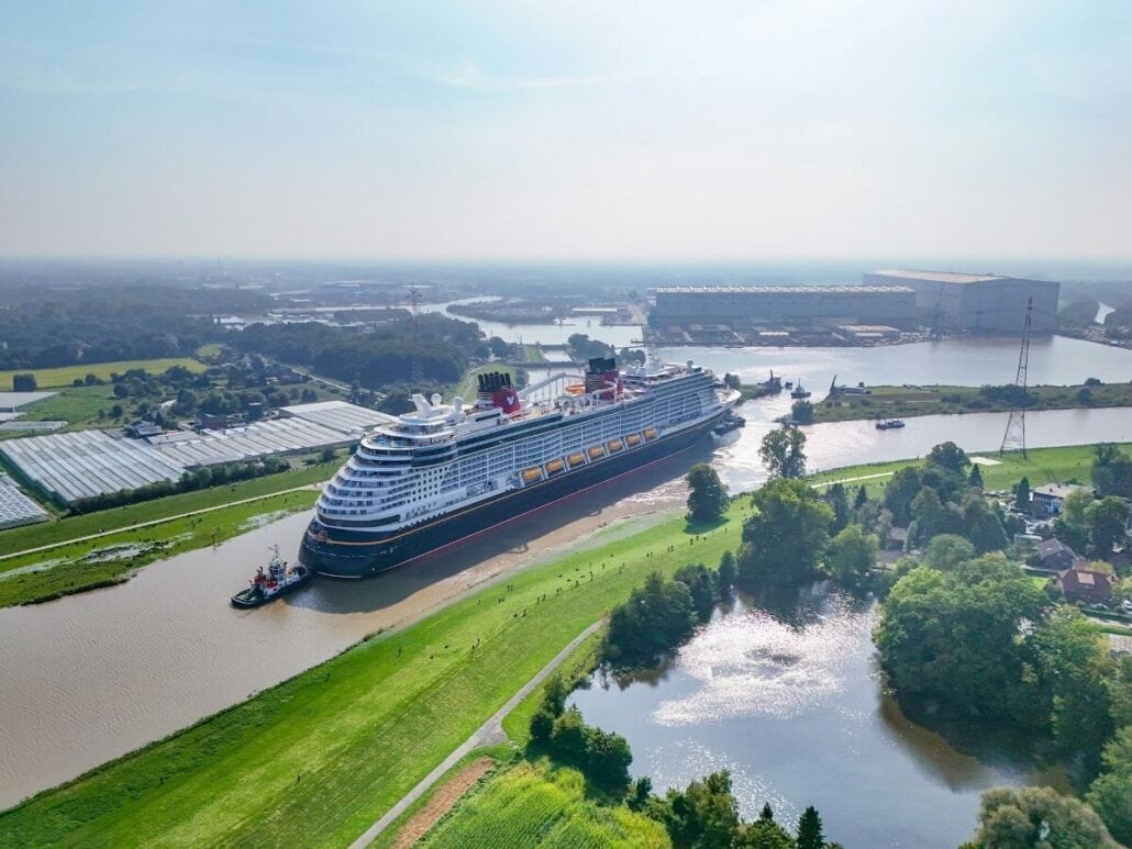 A large cruise ship, making its December debut, navigates through a narrow river surrounded by green fields and trees. The sky is clear, with several buildings in the background and a tugboat assisting this epic journey.