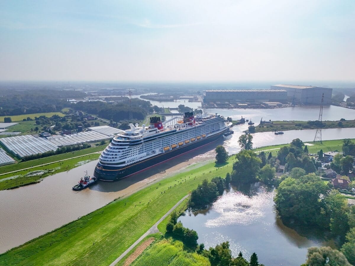 A large cruise ship, making its December debut, navigates through a narrow river surrounded by green fields and trees. The sky is clear, with several buildings in the background and a tugboat assisting this epic journey.
