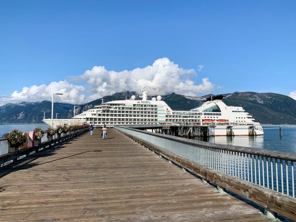 A majestic cruise ship is docked at a wooden pier, embraced by tranquil blue waters. Towering mountains and a partly cloudy sky form the stunning backdrop. Along the pier, adorned with flower baskets, strolls a couple enjoying the serene ambiance.