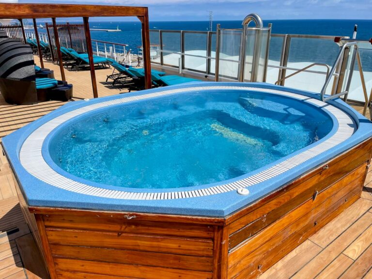 A hexagonal hot tub with blue water is built into a wooden deck. Lounge chairs are arranged nearby. The background shows a glass railing overlooking a body of water under a clear blue sky.