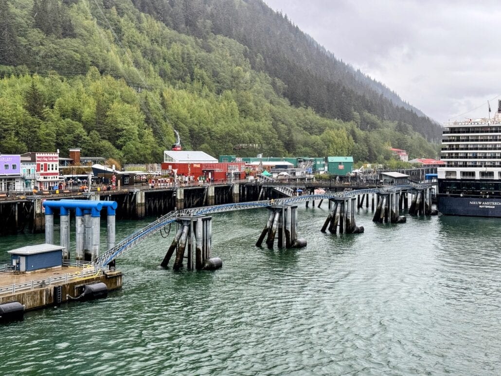 A waterfront scene on Ship-Free Saturday features a dock extending into the water, with colorful buildings nestled against a wooded hillside under a cloudy sky. Juneau voters enjoy the tranquility as no cruise ship is moored on the right, preserving the serene landscape.