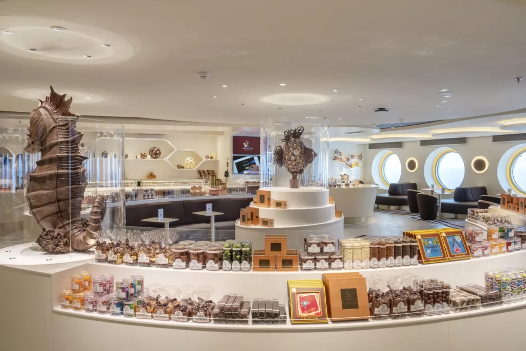 A modern chocolate shop interior features elegant displays of various chocolates and sweets. The room is well-lit with circular windows and decorative chocolate sculptures, including a large seahorse. Cozy seating resembling upscale bars is visible in the background.