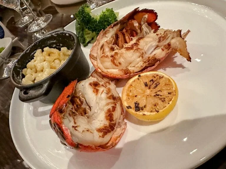 A plate with two halves of grilled lobster tail, a slice of grilled lemon, and a small pot of macaroni and cheese. There are broccoli florets placed on the plate as garnish.